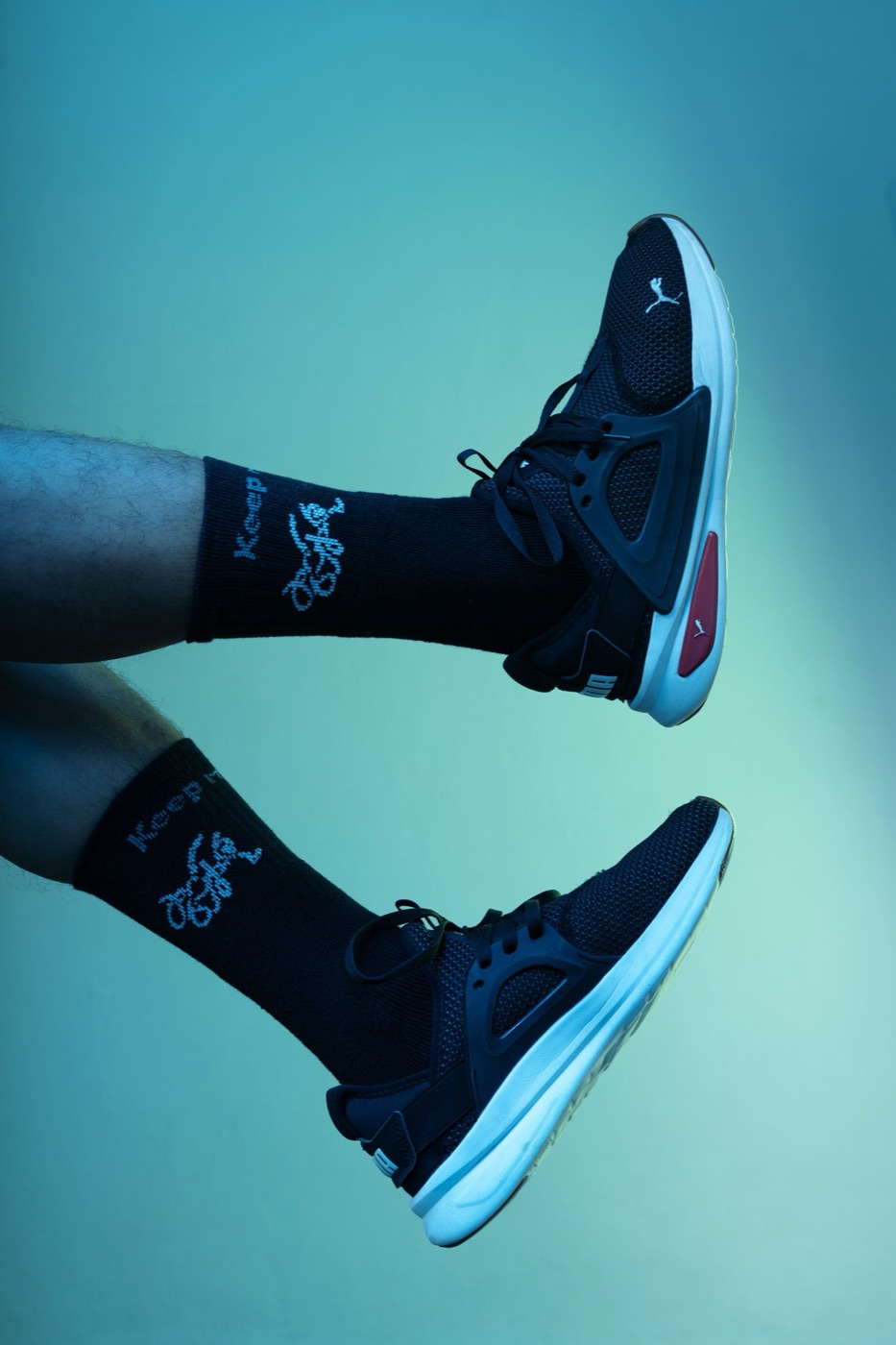 Legs kicked up in the air in black socks and sneakers against a teal backdrop.