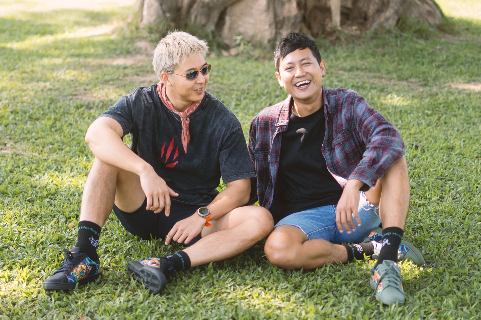 Two friends sitting on grass laughing, both wearing Keep Moving On socks and artwork sneakers.