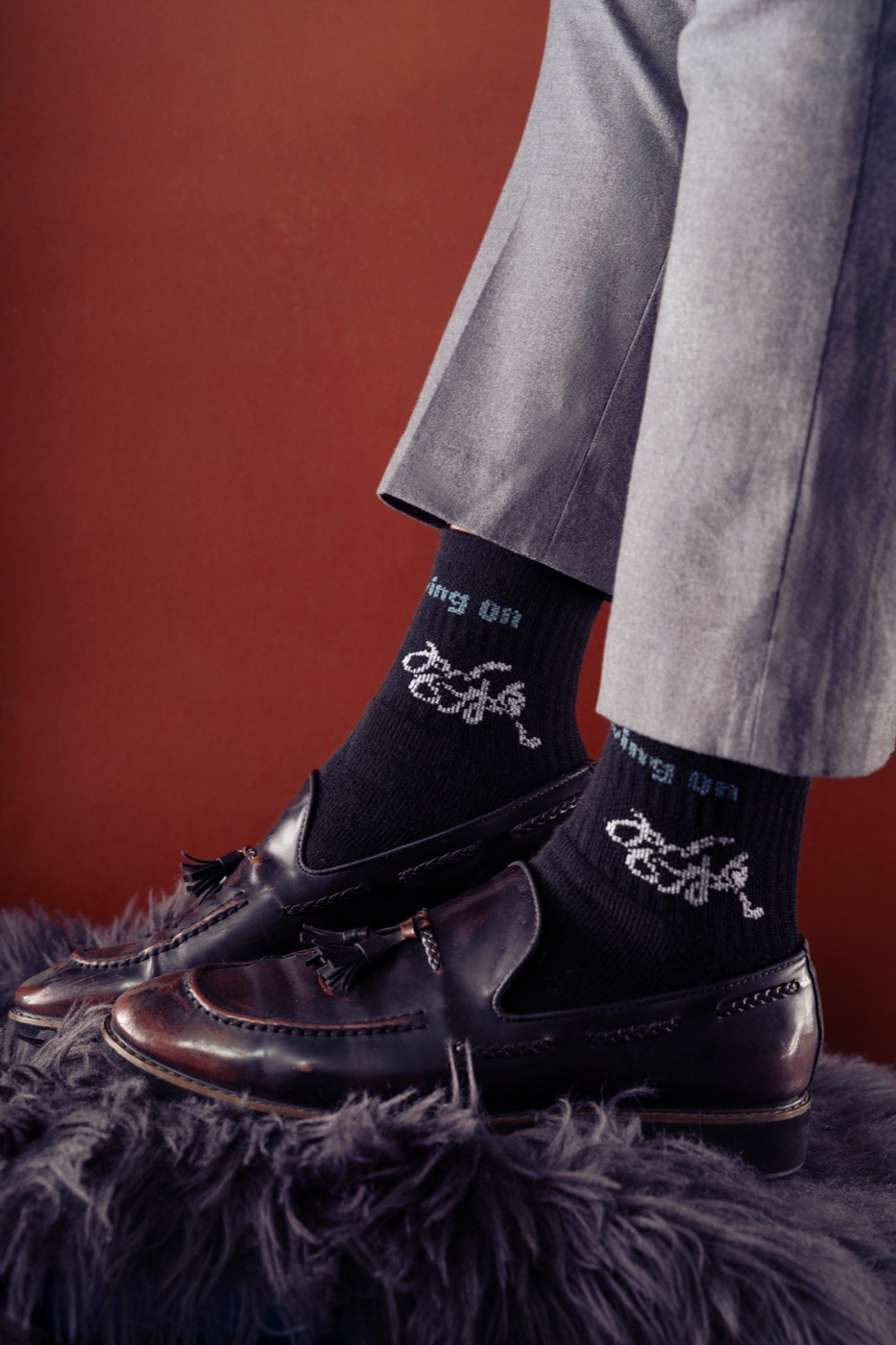 Side view of black crew socks and loafers on a fur stool against a red wall.