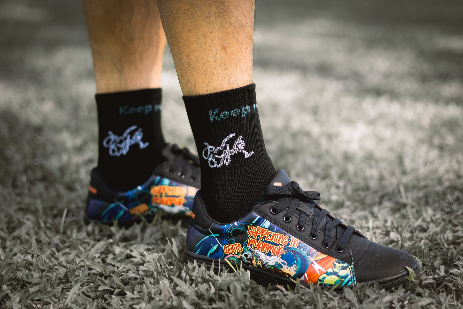 Sneakers painted with 'What's happening in Myanmar' artwork, worn with black Keep Moving On socks on grass.