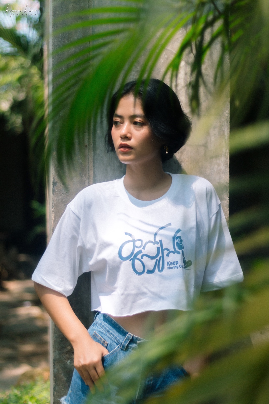 Model wearing the white Keep Moving On tee among palm leaves.