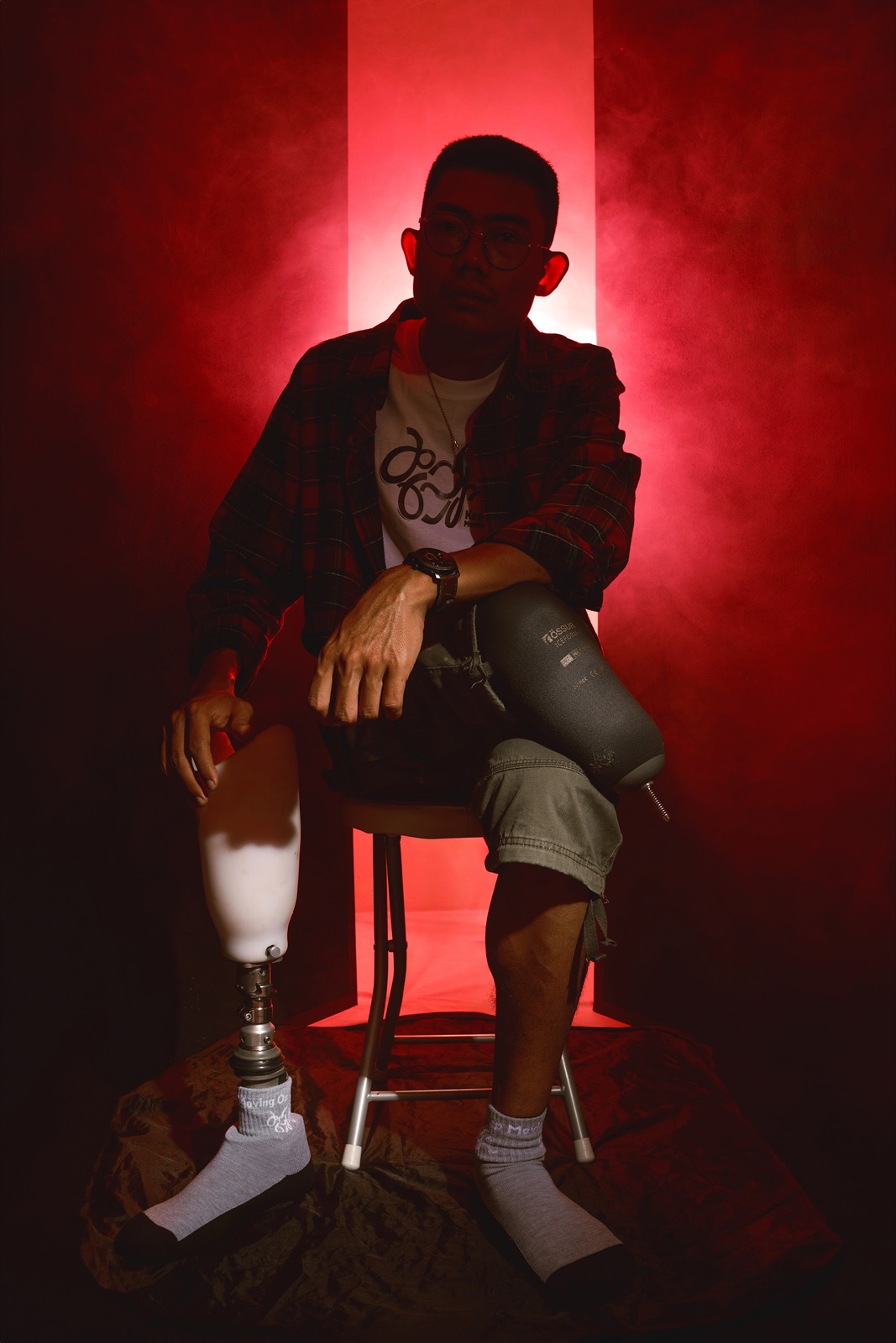 Portrait of a man seated in dramatic red light, one leg prosthetic, both feet wearing Keep Moving On socks.