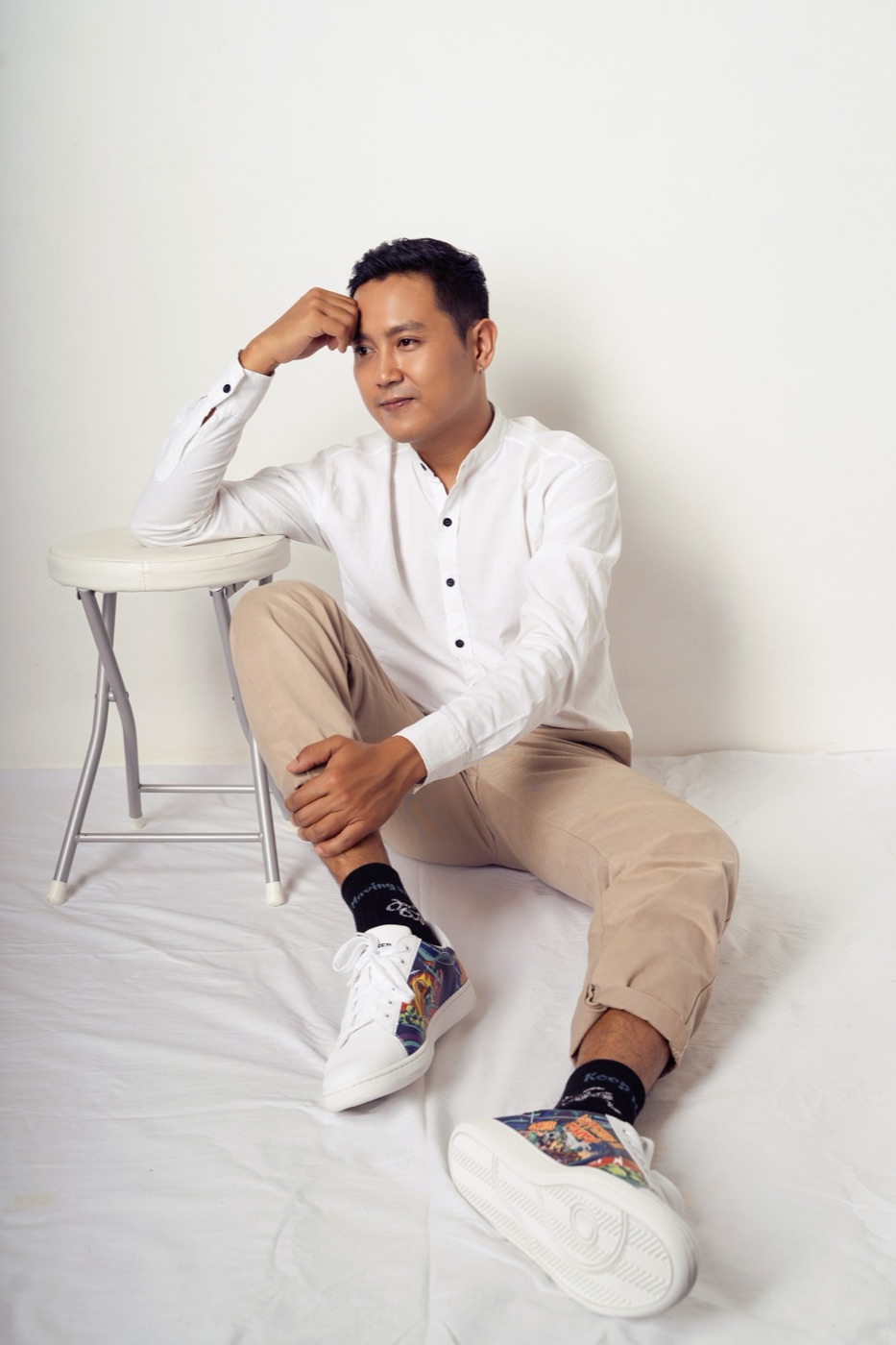 Man in a white shirt sitting on a studio floor wearing black Keep Moving On socks and artwork sneakers.