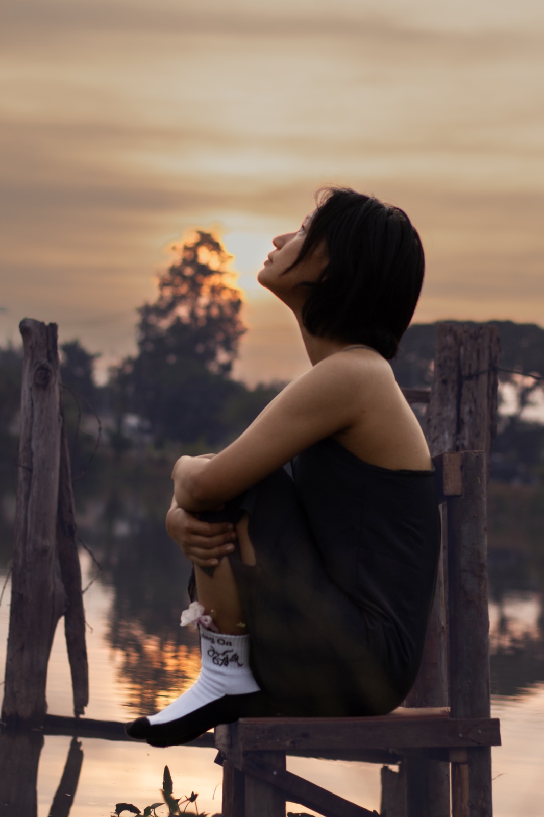 Woman in Keep Moving On socks sitting on a wooden chair above still water at golden hour, looking up at the sky.