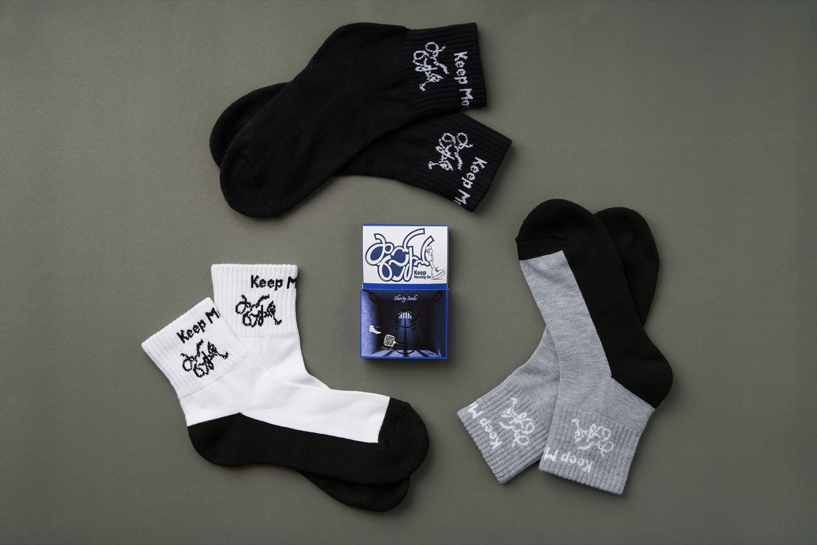 Flat lay of black, white and gray Keep Moving On socks around the packaging box.